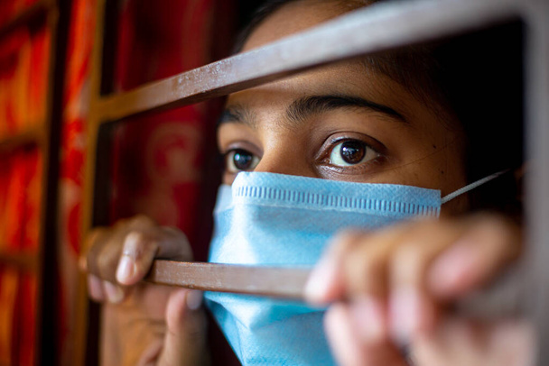 Znudzona Azjatka w masce chirurgicznej, patrząca przez okno na kwarantannę podczas epidemii koronawirusa. Widoki z bliska. - Zdjęcie, obraz