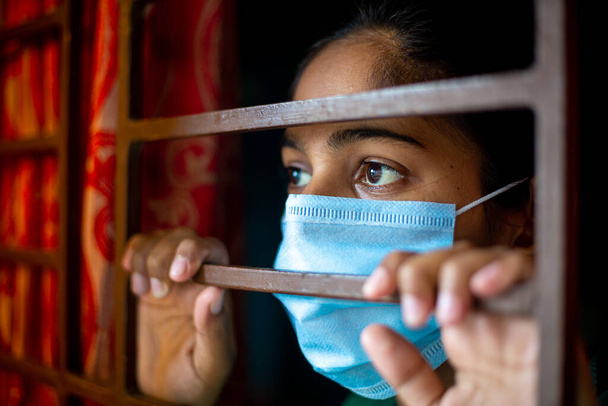 Ein gelangweiltes asiatisches junges Mädchen mit einer schützenden chirurgischen Gesichtsmaske, das während des Coronavirus-Ausbruchs durch ein Fenster in Quarantäne zu Hause schaut. Ansichten aus nächster Nähe. - Foto, Bild