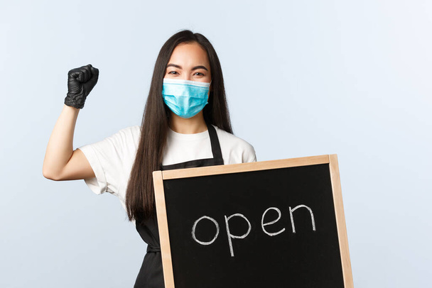 Small business, covid-19 pandemic, preventing virus and employees concept. Rejoicing cafe employee, store owner showing open sign and celebrating, wear medical mask and gloves - Фото, изображение