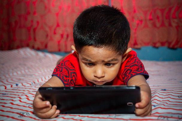 Un bambino con la massima attenzione sul suo letto a guardare cartoni animati utilizzando lo smartphone Tab. Bambini che giocano con lo smartphone. Concetto di telefonia mobile e dipendenza da Internet
. - Foto, immagini
