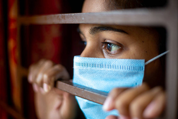 Une jeune fille asiatique ennuyée portant un masque chirurgical de protection à regarder par la fenêtre étant en quarantaine à la maison pendant l'épidémie de coronavirus. Vue rapprochée. - Photo, image