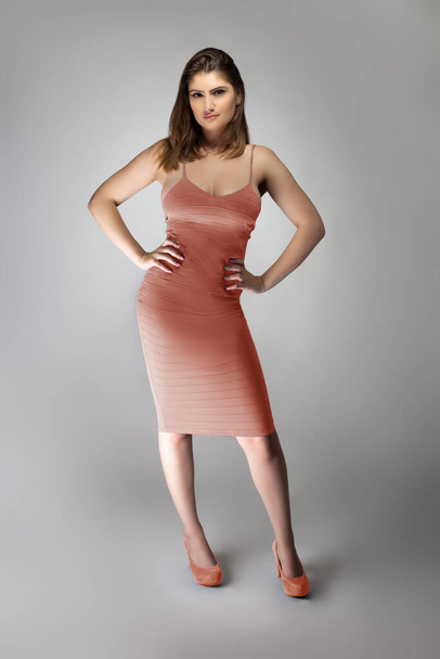 Confident female fashion model posing with a faded pink coral or flesh colored summer dress clothing in a studio for a catalog style portrait. The outfit is sleeveless and fit with matching shoes - Фото, изображение