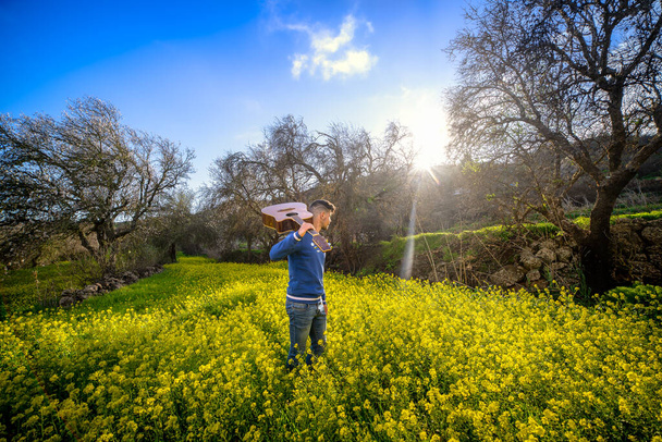 Omzunda gitar olan genç bir müzisyen, oturacak yer arıyor ve sarı çiçeklerle dolu bir tarlada oynuyor. - Fotoğraf, Görsel
