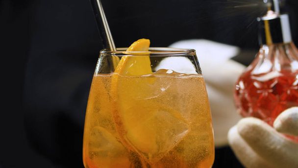 Aperol Spritz Cocktail σε ποτήρι κρασιού. Τα χέρια των μπάρμαν το πασπαλίζουν με αιθέριο έλαιο πορτοκαλιού. Μακρύ αφρόγαλα. - Φωτογραφία, εικόνα