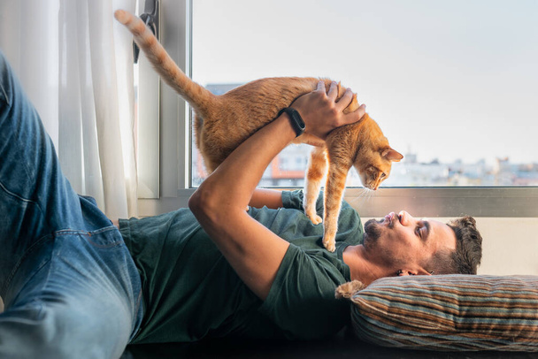 junger Mann, der unter dem Fenster liegt, nimmt eine gestromte Katze in den Arm und küsst sie vor sein Gesicht - Foto, Bild