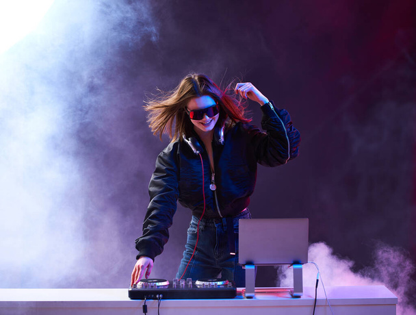 Stylish girl DJ in glasses headphones and a bomber mixes music dancing at a party in the neon light in the smoke . - Фото, изображение