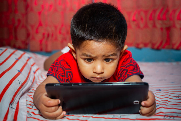 Een kind met volledige aandacht op zijn bed cartoons kijken met behulp van de Tab smartphone. Kinderen spelen met een smartphone. Mobiele telefoon en internetverslaving. - Foto, afbeelding