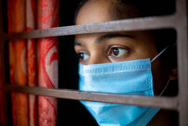 Una ragazza asiatica annoiata che indossa una maschera protettiva chirurgica a guardare attraverso la finestra in quarantena in casa durante l'epidemia di coronavirus. Osservazioni ravvicinate
. - Foto, immagini