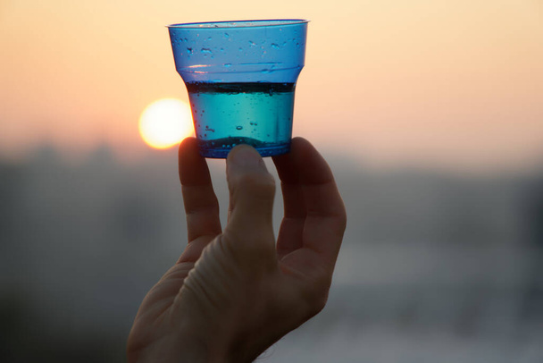 Man's hand holding glass against setting sun with blurry skyscrapers in cityscape background. - Photo, Image