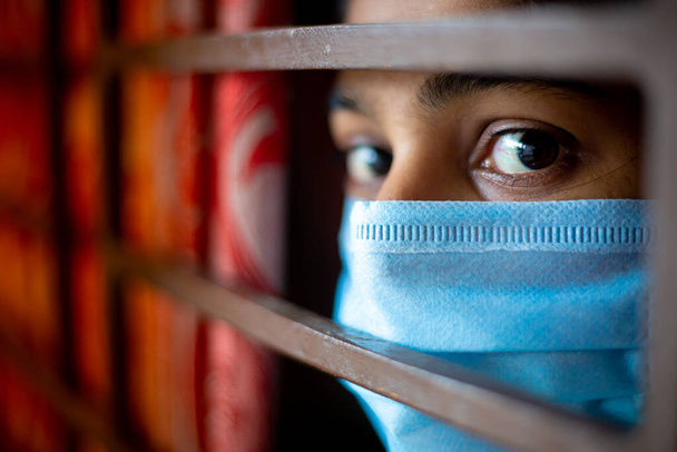 Una joven asiática aburrida que lleva una máscara quirúrgica de protección al mirar a través de la ventana estando en cuarentena en casa durante el brote de coronavirus. Vistas de cerca
. - Foto, imagen