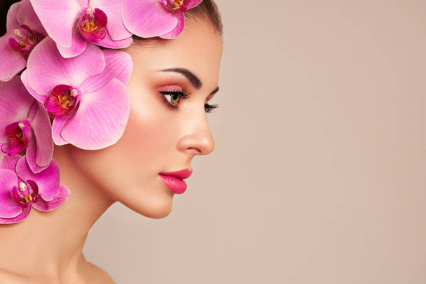 Portrait of beautiful young woman with orchid. Brunette woman with luxury makeup. Perfect skin. Eyelashes. Cosmetic eyeshadow. Purple flowers - Zdjęcie, obraz