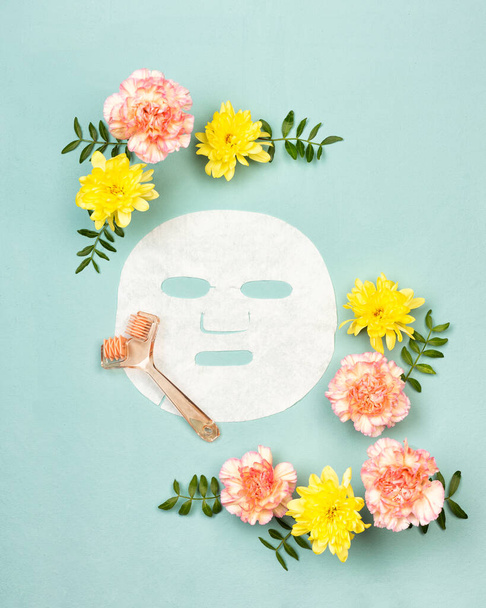 Face moistening sheet mask and massage roller in a frame of pink rose carnations and yellow chrysanthemums or mums flowers on pastel canvas blue background. Flat lay or top view, copy space for text - Photo, Image