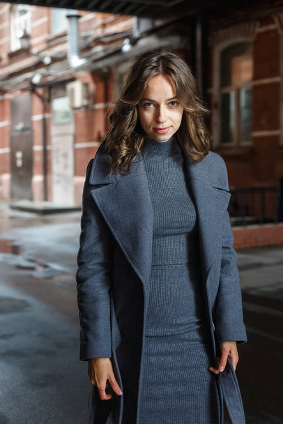 fiatal figyelmes csinos lány szürke kabátban és garbóban pózol a vörös épület előtt. Street style portré fotó ülésen vonzó nő, városi fotózás gyönyörű elegáns modell - Fotó, kép