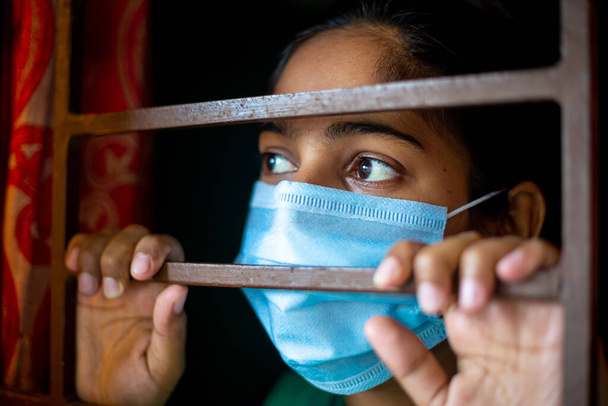 Ένα νεαρό κορίτσι από την Ασία που βαριέται και φοράει μάσκα προστασίας κοιτάζοντας μέσα από το παράθυρο να είναι σε καραντίνα στο σπίτι κατά τη διάρκεια της επιδημίας του κορωνοϊού. Κοντινές προβολές. - Φωτογραφία, εικόνα
