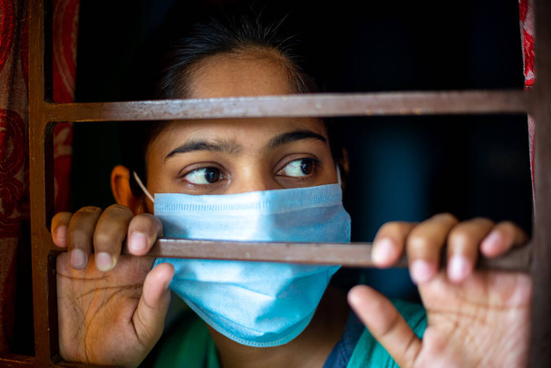 Una joven asiática aburrida que lleva una máscara quirúrgica de protección al mirar a través de la ventana estando en cuarentena en casa durante el brote de coronavirus. Vistas de cerca
. - Foto, Imagen