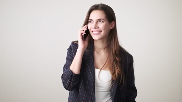 A smiling cheerful young woman wearing casual clothes is talking on the phone standing isolated over white background in studio - Materiał filmowy, wideo