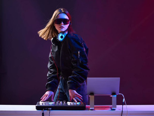 Stylish DJ girl with headphones and glasses on the bomber jacket mixes the music at the party and neon lights. - Fotografie, Obrázek