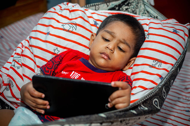 Antes de dormir, uma criança está deitada em uma rede caseira e assistindo desenhos animados usando uma aba do smartphone. Crianças brincando com smartphone. Conceito de vício em celular e internet
. - Foto, Imagem