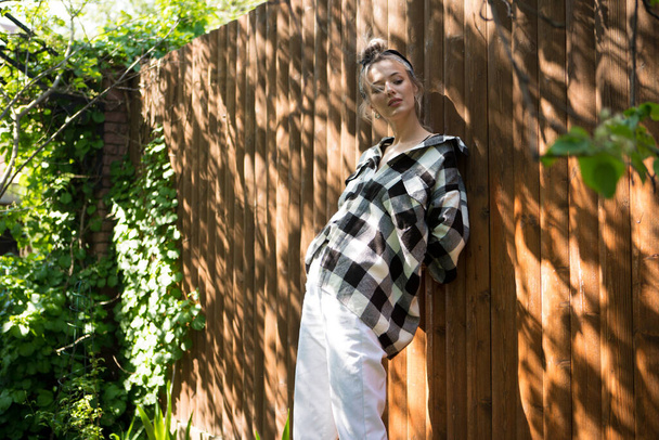 Portrait of a pretty girl in a plaid shirt and bandana standing against a wooden fence in the backyard on a Sunny day. - Foto, Imagem
