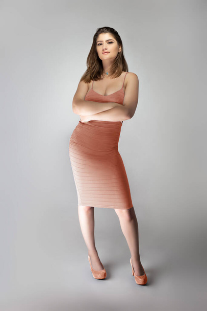 Confident female fashion model posing with a faded pink coral or flesh colored summer dress clothing in a studio for a catalog style portrait. The outfit is sleeveless and fit with matching shoes - Zdjęcie, obraz