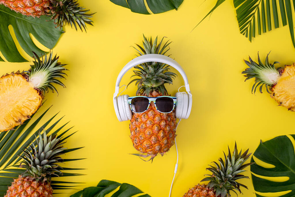 Funny pineapple wearing white headphone, listen music, isolated on yellow background with tropical palm leaves, top view, flat lay design concept. - Φωτογραφία, εικόνα