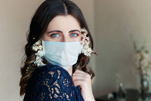 Junge hübsche Frau in medizinischer Maske, geschmückt mit Frühlingsblumen, bleibt zu Hause zur Selbstquarantäne isoliert. Konzept Heimquarantäne, Prävention COVID-19, Coronavirus-Ausbruchssituation. - Foto, Bild