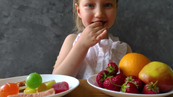 Κοριτσάκι με φρέσκα φρούτα και γλυκά στο τραπέζι στο σπίτι. Το παιδί σκέφτεται τι να επιλέξει υγιεινό ή επιβλαβές πρόχειρο φαγητό. Παιδί επιλέγει και απολαμβάνει να τρώει φυσικά μούρα το καλοκαίρι, βιταμίνες χορτοφαγικό γεύμα. - Πλάνα, βίντεο