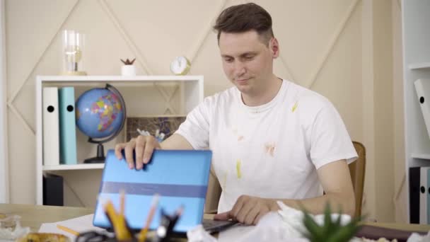 Portrait of satisfied young man closing laptop and leaning back on chair. Brunette Caucasian male freelancer in dirty T-shirt ending online project. Freelance, lifestyle, remote work, happiness. - Кадри, відео