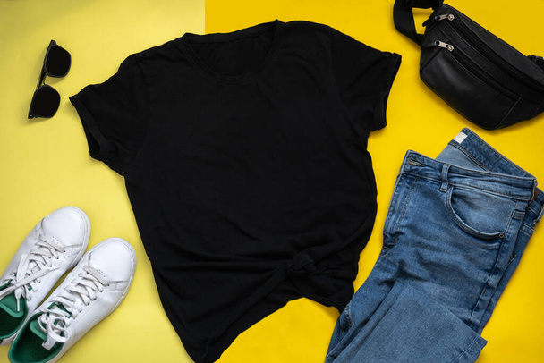 Черный макет футболки на жёлтом фоне. Квартира с хипстерской одеждой. Голубые джинсы, черные солнечные очки, белые кроссовки и плечевая сумка
 - Фото, изображение
