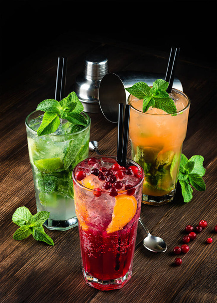 three alcoholic cocktails mojito and cranberry or lingonberry cocktail on a wooden background, refreshing mojito cocktail with ice and mint in a glass. cocktail decorated with cranberries - Φωτογραφία, εικόνα