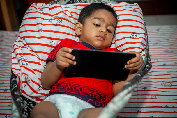 Vor dem Schlafengehen liegt ein Kind in einer selbstgemachten Hängematte und schaut sich Cartoons per Smartphone-Tab an. Kinder spielen mit dem Smartphone. Suchtkonzept für Mobiltelefone und Internet. - Foto, Bild