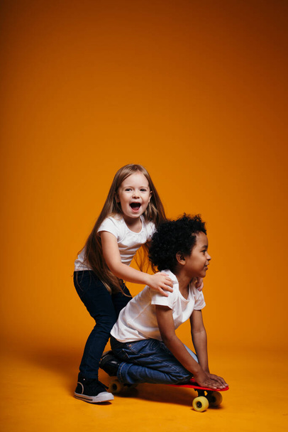 The girl laughs loudly while rolling her black friend on a longboard on an orange background in the studio - Foto, Imagem
