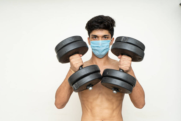 Фитнес-человек в маске для защиты от эпидемии Рабочие упражнения для рук, бицепсов и плеч с гантелями.
 - Фото, изображение