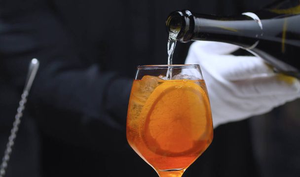 Preparing an Aperol Spritz Cocktail. Close up bartenders hands pouring prosecco in wine glass. Long fizzy drink. - Photo, Image