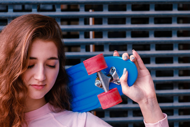 Ginger girl posing against a metal mesh with her eyes closed and holding her blue skateboard on her shoulder. - 写真・画像