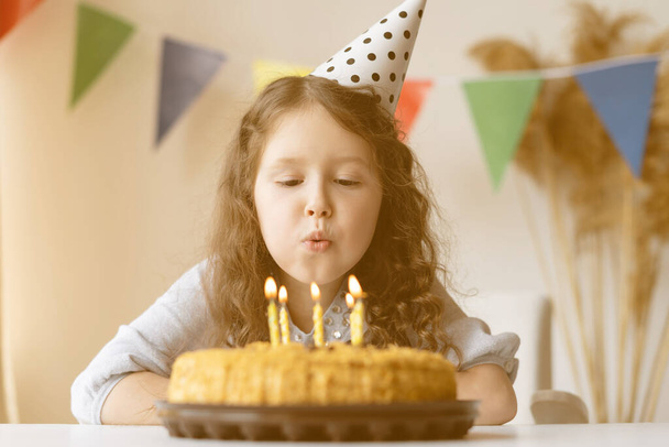 Little blonde girl smiling in birthday pink cap, a chocolate birthday cake with candles. Child celebrates her birthday. Happy birthday. - Photo, image