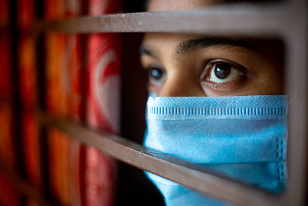 Скучающая азиатская молодая девушка в защитной хирургической маске, смотрящая в окно, будучи в домашнем карантине во время вспышки коронавируса. Крупный план
. - Фото, изображение