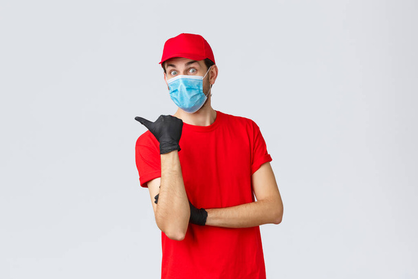 Covid-19, self-quarantine, online shopping and shipping concept. Excited surprised delivery man in red uniform, face mask and gloves, pointing left at promo, showing clients bonuses or application - Photo, Image