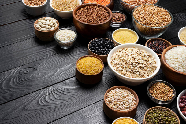 Various cereals, grains, seeds, groats, legumes and beans in bowls on black wooden background with copy space - Photo, Image