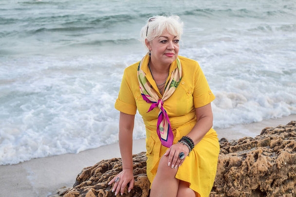 όμορφη ενήλικη γυναίκα με χτένισμα και μακιγιάζ σε κίτρινο φόρεμα ποζάρουν στην παραλία  - Φωτογραφία, εικόνα