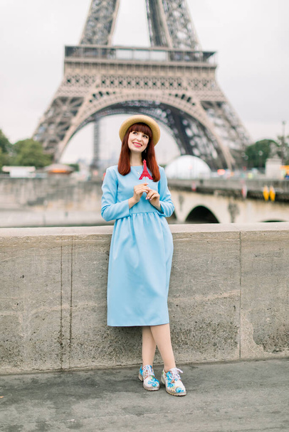 pretty young woman in blue dress and straw hat with red eiffel tour shaped lollipop in hands standing on the background of Eiffel tower in Paris - Фото, зображення