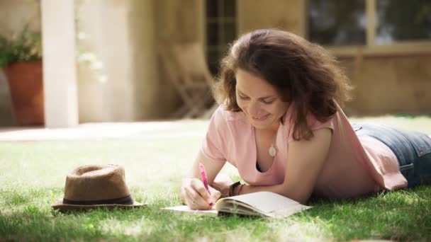  Nuori onnellinen nainen makaa nurmikolla maalaistalossa ja tekee muistiinpanoja muistikirjaan - Materiaali, video