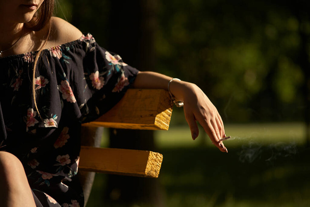 Крупный план женской руки с сигаретой в парке. Концепция курения табака в общественном месте, вредного для здоровья и других, наркомании и рака
. - Фото, изображение