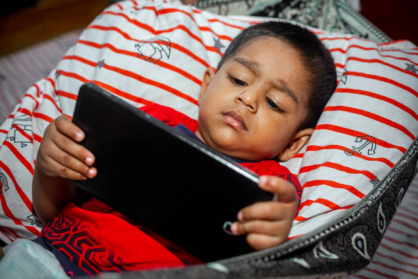 Перед сном ребенок лежит домашний гамак и смотрит мультики, используя вкладку смартфона. Дети играют со смартфоном. Концепция мобильного телефона и интернет-зависимости
. - Фото, изображение