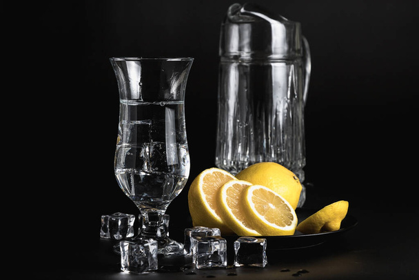 Still life on a black background. Glass and ice. Cold drinking water. Yellow lemon in a black plate. - Фото, изображение