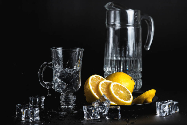 Still life on a black background. Glass and ice. Cold drinking water. Yellow lemon in a black plate. Ice falls into the glass. A spray of water. - Photo, image