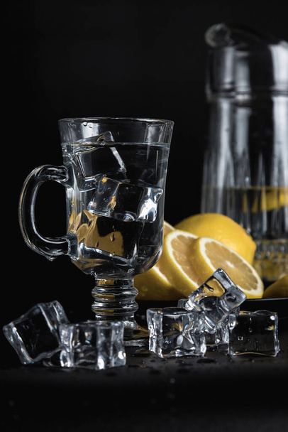 Sliced lemon on a black plate. A decanter and a tall glass of water and ice. Still life of glass objects on a black background. Cold lemonade. Yellow on black. - Photo, image