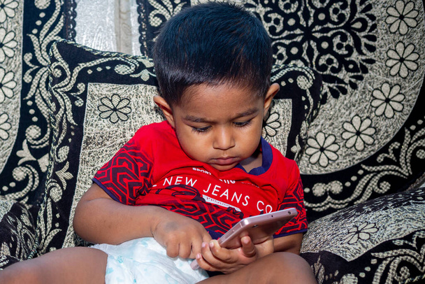 Egy ázsiai kis 18 hónapos kisfiú ül a kanapén és egy videót néz a Tab okostelefonról. A gyerekek okostelefonnal játszanak. Mobiltelefon és internet-függőség fogalma. - Fotó, kép