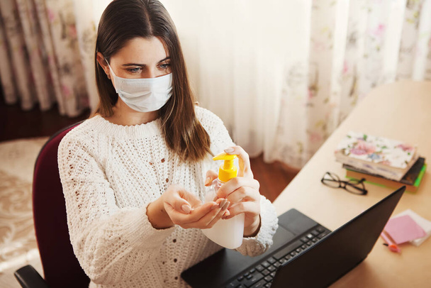 Coronavirus concept. Woman in medical mask working from home. Business woman at home quarantine wearing protective mask. Working from home. Cleaning her hands with sanitizer gel - Photo, Image