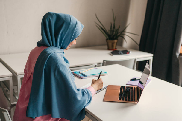 young pretty modern muslim woman in hijab working on laptop in office room, education online, remote work freelancer, making notes thinking - Foto, Imagen